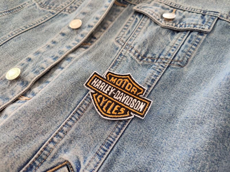 Fotografia di un dettaglio di un giubbotto di jeans con toppa a forma del logo Harley-Davidson Motorcycles arancione bianca e nera