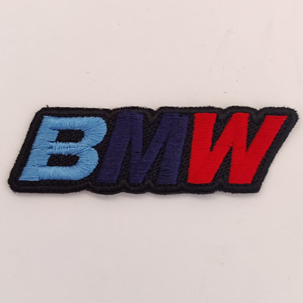 Toppa ricamata con base nera e con lettera B in azzurro, la lettera M in blu scuro e la lettera W in rosso