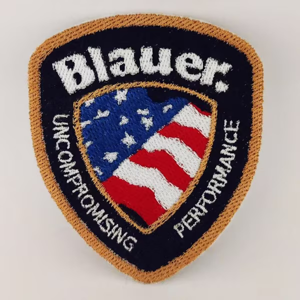 Fotografia della toppa ricamata Blauer a forma di scudetto nera, con contorni oro, al centro la bandiera degli Stati Uniti d'America e le scritte bianche.