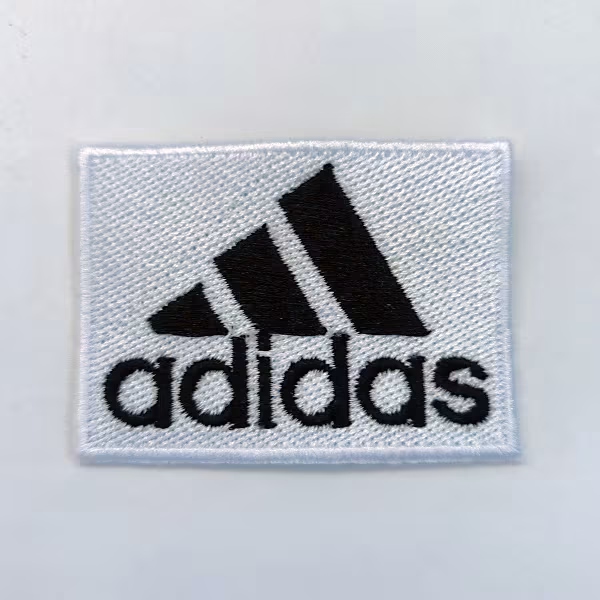 Toppa rettangolare bianca con logo nero Adidas e tre strisce nere su sfondo bianco.