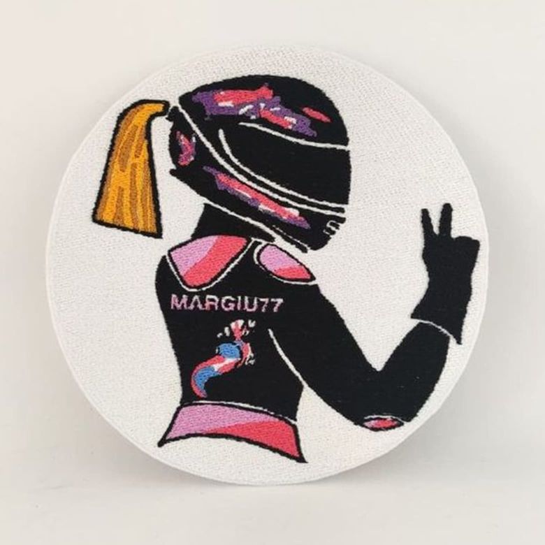 Fotografia di una toppa circolare bianca con dentro disegnata una motociclista donna con casco e coda di cavallo bionda