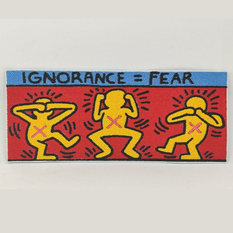Fotografia della toppa ricamata rettangolare dell'opera dell'artista Keith Haring con tre omini gialli con croci rosa al centro su sfondo rosso e blu con scritto in alto "Ignorance = Fear"