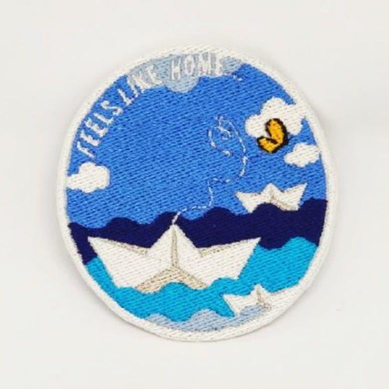 Toppa ricamata circolare di una barca di carta in mare con scritta "Feels Like Home"